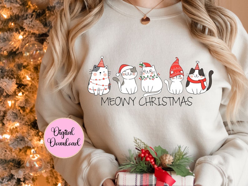 May include: Cream-coloured sweatshirt with a Christmas-themed design of five cartoon cats wearing festive hats and scarves. The words "MEOWY CHRISTMAS" are written below the cats. A pink circle with the words "Digital Download" is in the lower left corner.