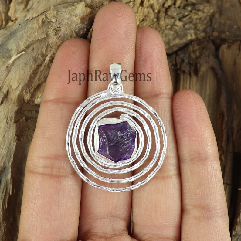 May include: A silver pendant with a spiral design and a purple amethyst gemstone in the center. The pendant is on a hand, and the text "JaphRawGems" is visible in the background.