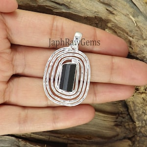 May include: Silver pendant with a black tourmaline gemstone set in a circular design. The pendant is surrounded by a textured silver frame.