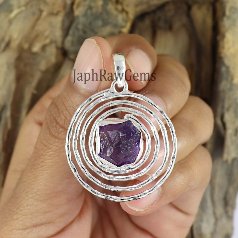 May include: A silver pendant with a spiral design and a purple amethyst gemstone in the center. "JaphrRawGems" is written on the pendant.