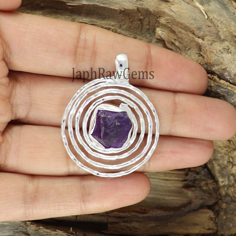 May include: A silver pendant with a spiral design and a purple amethyst gemstone in the center. The pendant is on a hand.