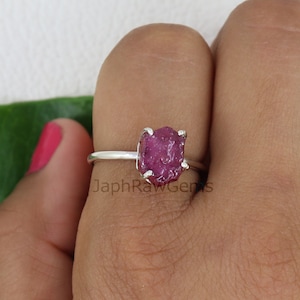 Op de afbeelding: Een zilveren ring met een ruwe, roze robijn edelsteen. De ring wordt gedragen aan een vinger.