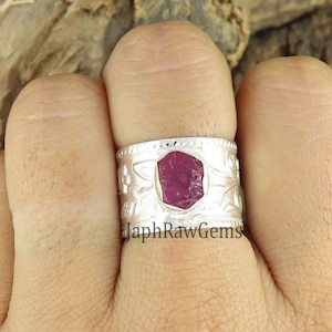 Może przedstawiać: Srebrny pierścionek z dużym, surowym rubinem osadzonym w środku. Obrączka ozdobiona jest misternymi wzorami kwiatowymi.