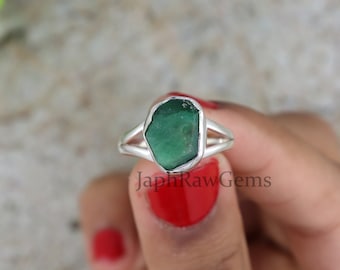 Può includere: Un anello d'argento con una pietra preziosa di smeraldo verde grezzo. L'anello ha una montatura a doppia fascia.