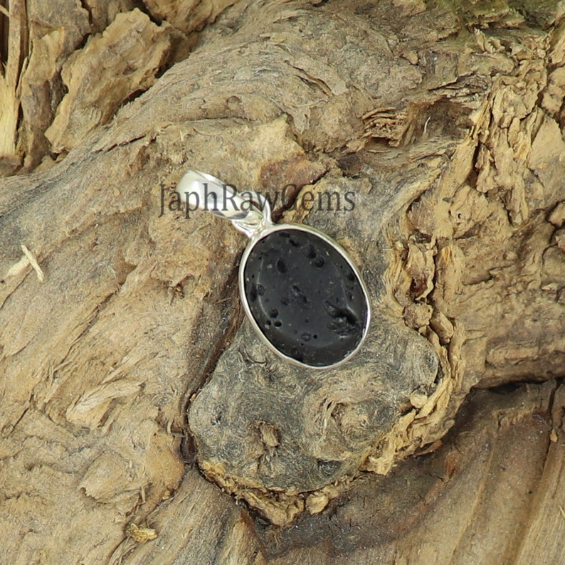 May include: A black oval lava rock pendant with a silver bail. The pendant is sitting on a piece of wood.
