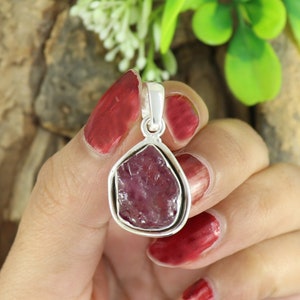 Puede incluir: Un colgante de plata con una piedra preciosa de rubí rosa en bruto engastada en un engaste de plata. El colgante se sostiene en una mano con esmalte de uñas rojo.