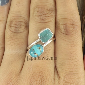 Puede incluir: Un anillo de plata con dos piedras turquesa. La piedra m&aacute;s grande es de forma ovalada y la m&aacute;s peque&ntilde;a es de forma cuadrada. El anillo est&aacute; en un dedo.