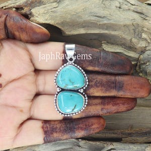 May include: A silver pendant with two turquoise-colored stones. The stones are oval-shaped and set in a silver frame with a beaded edge. The pendant is held in a hand, with the text "JaphRawGems" visible.