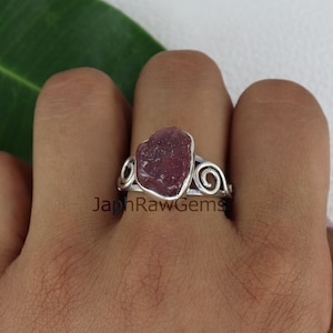 May include: A silver ring with a deep red, raw ruby gemstone set in a delicate, swirling design.  The ring is on a person's finger.