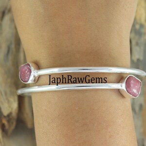 May include: A silver bangle bracelet with two pink gemstones. The bracelet is on a person's wrist. The text "JaphRawGems" is visible on the bracelet.