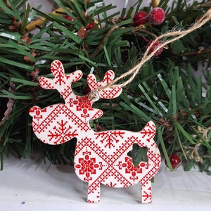Puede incluir: Adorno navideño de reno de madera con un diseño estampado en rojo y blanco. El adorno tiene un recorte en forma de estrella y está suspendido por una cuerda de hilo natural. El reno se encuentra sobre un fondo de agujas de pino verdes y bayas rojas.