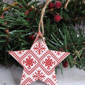 Puede incluir: Adorno navideño en forma de estrella de madera con un diseño nórdico rojo y blanco. Cuelga de una cuerda de hilo y tiene una pequeña campana plateada. El fondo presenta una guirnalda verde con bayas rojas, creando un ambiente festivo.