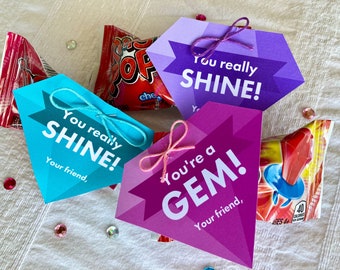 Può includere: Tre carte a forma di diamante con messaggi incoraggianti: "You really SHINE!" in turchese, "You really SHINE!" in viola e "You're a GEM!" in rosa. Ogni carta è legata con un nastro e posizionata sopra un piccolo sacchetto di caramelle.