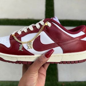May include: White and red leather low-top sneakers with a gold chain and "LA" charms. The chain is attached to the laces and hangs down the side of the shoe.