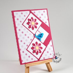 May include: A pink card with a white background and a pink and yellow flower design. The card is folded in half and has a blue butterfly on the front. The card is sitting on a wooden easel.