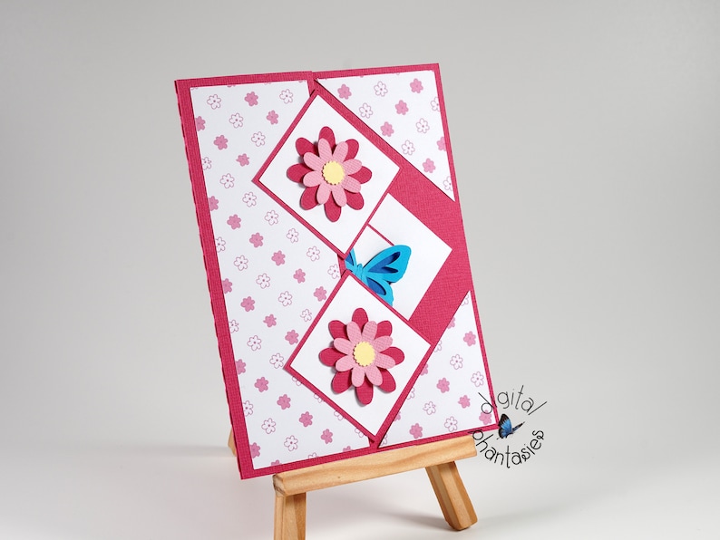 May include: A pink card with a white background and a pink and yellow flower design. The card is folded in half and has a blue butterfly on the front. The card is sitting on a wooden easel.