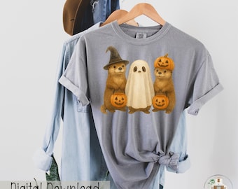 May include: A gray t-shirt featuring a Halloween design with three otters. One otter wears a witch hat, another is dressed as a ghost, and the third wears a pumpkin on its head. Each otter holds a jack-o'-lantern. The words "Digital Download" are visible.