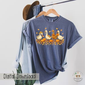 May include: A blue t-shirt featuring a fall-themed design with four cartoon geese wearing hats and scarves, standing among pumpkins and autumn leaves. The words "Digital Download" are printed on the bottom left.