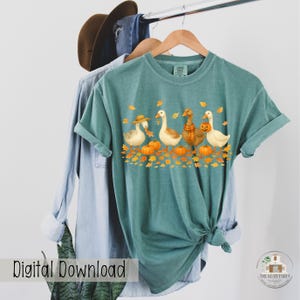 May include: Teal-colored t-shirt featuring a fall-themed graphic. The design includes five geese, pumpkins, and autumn leaves. One goose wears a hat, another a scarf, and one carries a pumpkin. The words "Digital Download" are visible on the bottom left.