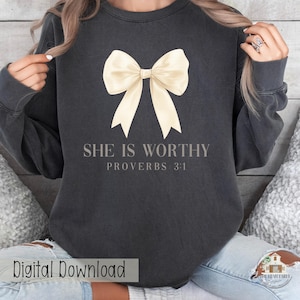 May include: Dark gray sweatshirt with a cream-colored bow graphic. The text "SHE IS WORTHY PROVERBS 3:1" is printed below the bow. The words "Digital Download" are printed at the bottom left.