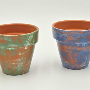 May include: Two terracotta plant pots with a distressed finish. The left pot features a green and silver brushed texture over the natural clay, while the right pot displays a blue and silver brushed texture over the same terracotta base.