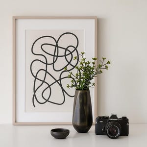 Puede incluir: Una impresión artística abstracta enmarcada con líneas negras sobre un fondo crema. Un jarrón oscuro contiene vegetación, junto a un pequeño cuenco negro y una cámara negra vintage. Los objetos están dispuestos sobre una superficie blanca.