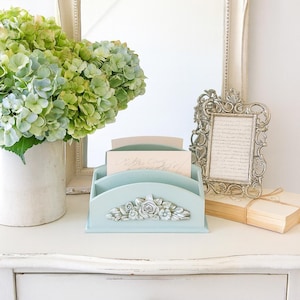 Op de afbeelding: Een lichtblauwe bureau-organizer met een bloemenontwerp, met briefpapier. Een boeket groene hortensia's in een witte vaas, een ingelijste spiegel en een decoratief frame staan ook op het witte oppervlak.