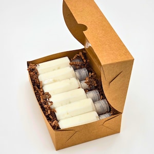 Könnte beinhalten: Eine braune Pappschachtel mit teilweise geöffnetem Deckel, gefüllt mit mehreren transparenten Plastikröhrchen, die eine weiße Substanz enthalten. Die Röhrchen sind ordentlich auf einem Bett aus braunem Zierpapier angeordnet.