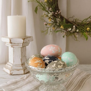 Könnte beinhalten: Eine Glasschale gefüllt mit dekorierten Ostereiern in Pastelltönen, darunter Rosa, Mintgrün und Orange, mit floralen Mustern. Eine weiße Säulenkerze steht auf einem weißen, antiken Sockel. Ein dekorativer Kranz hängt im Hintergrund.
