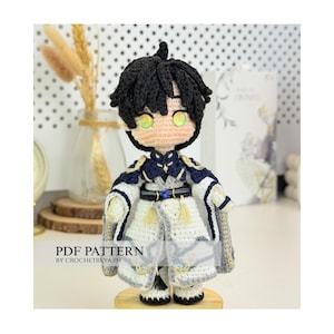 Op de afbeelding: Een handgemaakte gehaakte pop met zwart haar en groene ogen, gekleed in een wit en marineblauw outfit met gouden accenten. De pop staat op een houten voet. De tekst "PDF PATTERN BY CROCHETREYA.PH" is zichtbaar.