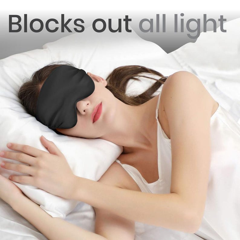 May include: A person wearing a black sleep mask while lying in bed. The mask covers their eyes completely. The text "Blocks out all light" is visible at the top of the image.