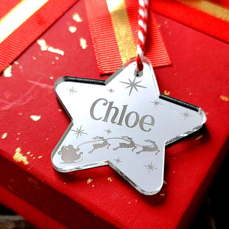 May include: A star-shaped Christmas ornament with the name "Chloe" engraved on it. The ornament is silver and features a design of Santa's sleigh and reindeer. It hangs from a red and white string, resting on a red gift box with a red ribbon.