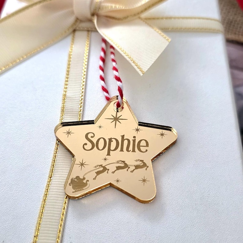 May include: A gold star-shaped gift tag with the name "Sophie" engraved on it. The tag features a design of Santa Claus in a sleigh pulled by reindeer. The tag hangs from a red and white string, and is attached to a white gift box with a cream ribbon.