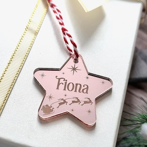 May include: A pink star-shaped Christmas ornament with the name "Fiona" engraved on it. The ornament features a sleigh and reindeer design, with a red and white string for hanging. Decorative stars are also engraved on the ornament.