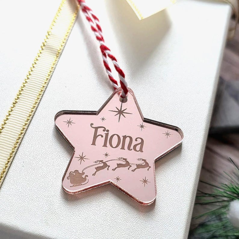 May include: A pink star-shaped Christmas ornament with the name "Fiona" engraved on it. The ornament features a sleigh and reindeer design, with a red and white string for hanging. Decorative stars are also engraved on the ornament.