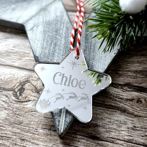 May include: A mirrored star-shaped Christmas ornament with the name "Chloe" engraved. The ornament features a red and white striped string for hanging and a festive design of reindeer and a sleigh. The background includes greenery and a wooden surface.