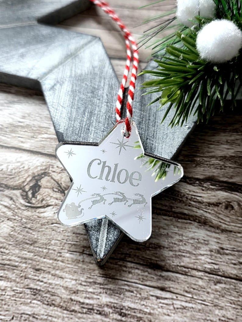 May include: A mirrored star-shaped Christmas ornament with the name "Chloe" engraved. The ornament features a red and white striped string for hanging and a festive design of reindeer and a sleigh. The background includes greenery and a wooden surface.