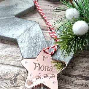 May include: A rose gold star-shaped Christmas ornament engraved with the name "Fiona" and a Santa and reindeer design. The ornament hangs from a red and white striped cord. Decorative greenery and a wooden star are in the background.