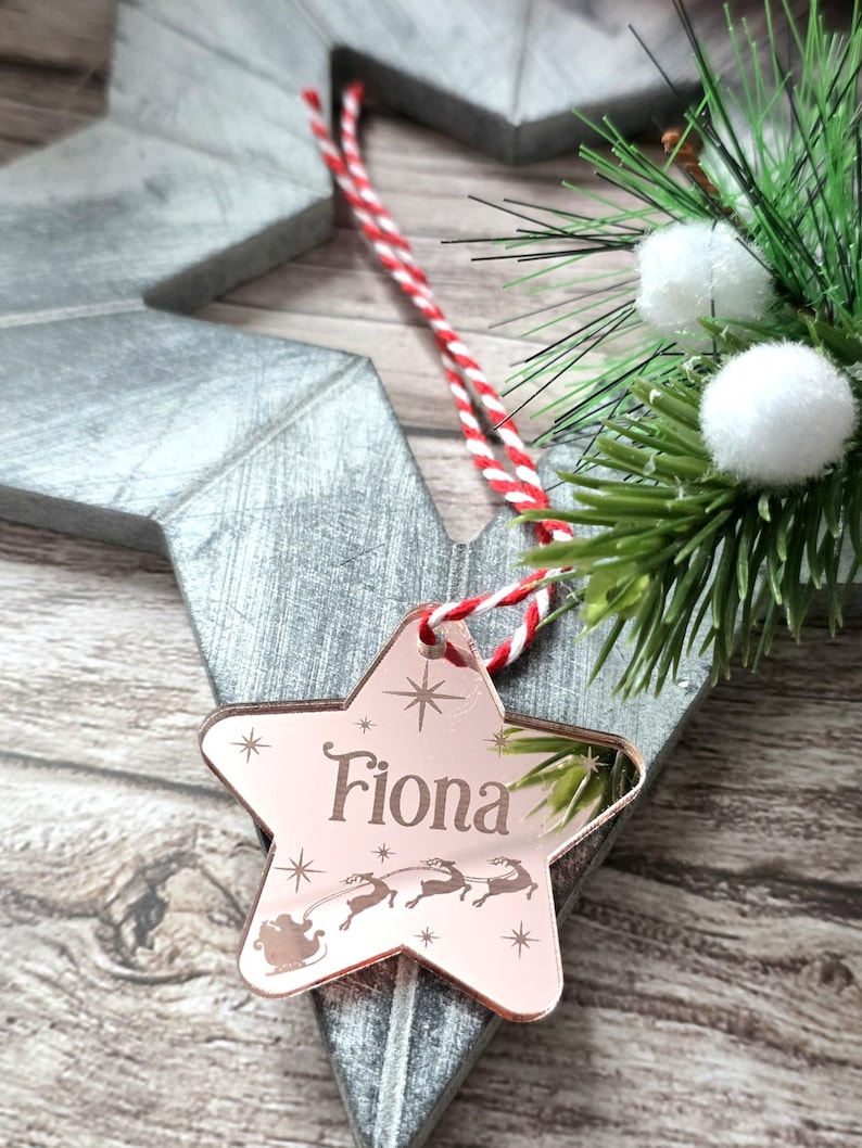 May include: A rose gold star-shaped Christmas ornament engraved with the name "Fiona" and a Santa and reindeer design. The ornament hangs from a red and white striped cord. Decorative greenery and a wooden star are in the background.