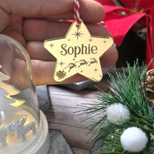 May include: A gold star-shaped Christmas ornament with the name "Sophie" engraved on it. The ornament features a design of Santa's sleigh and reindeer. It hangs from a red and white string, perfect for holiday decor.