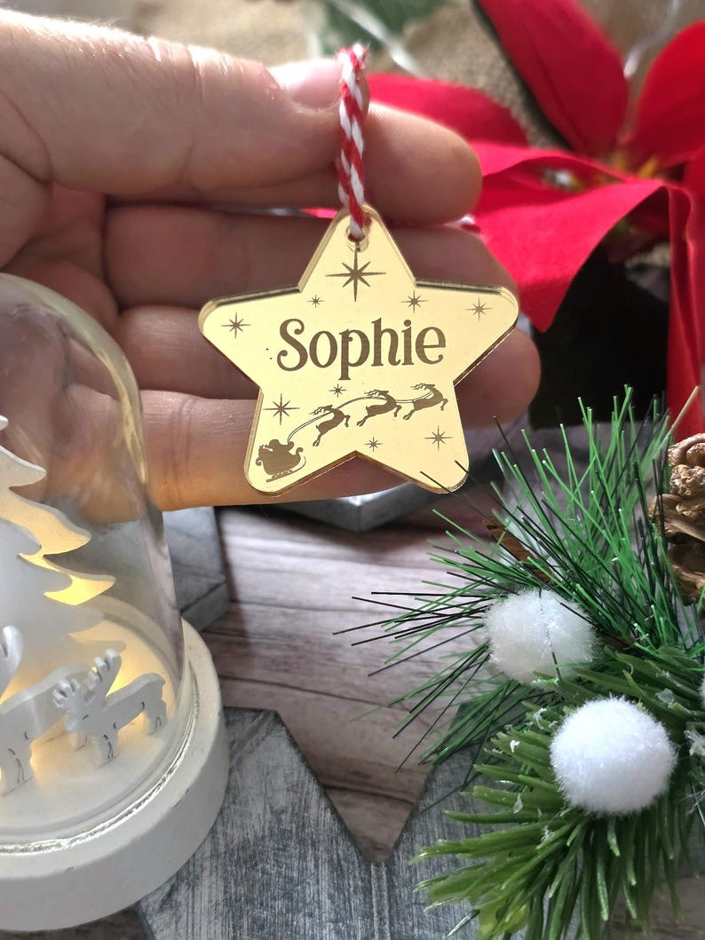 May include: A gold star-shaped Christmas ornament with the name "Sophie" engraved on it. The ornament features a design of Santa's sleigh and reindeer. It hangs from a red and white string, perfect for holiday decor.