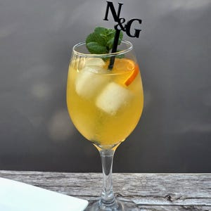 May include: A glass of yellow cocktail with ice cubes and a slice of orange. A black plastic stirrer with the letters "N & G" is in the drink.