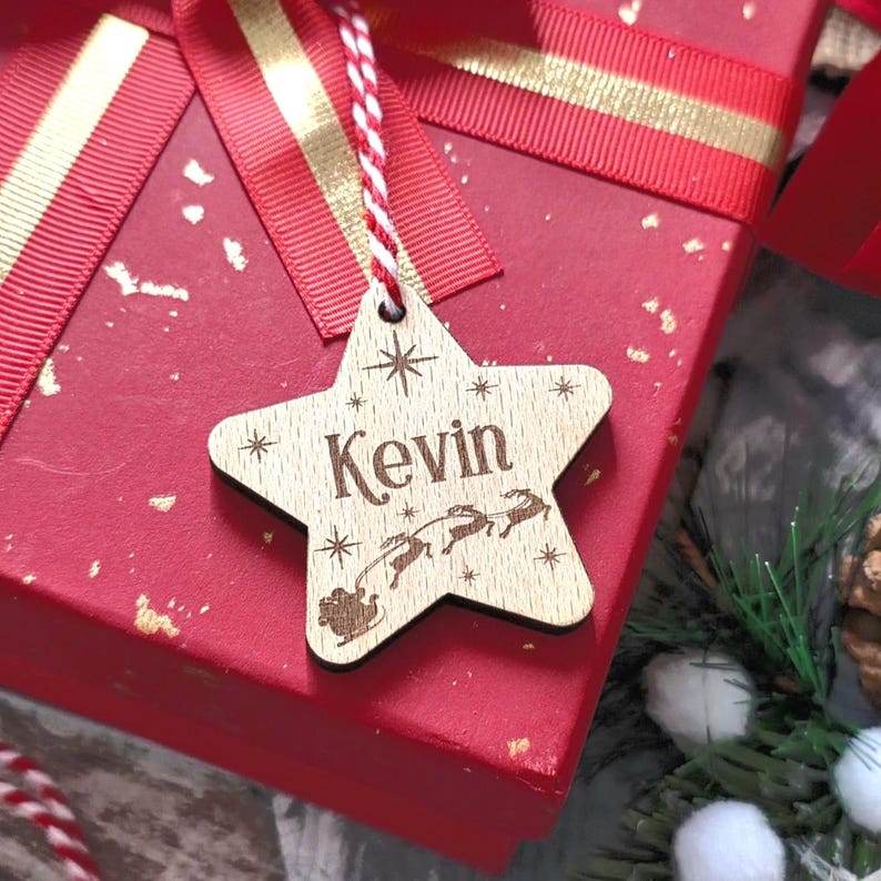 May include: A wooden star-shaped gift tag with the name "Kevin" engraved on it. The tag features a Santa Claus and reindeer design. The tag is hanging from a red and white string, and is placed on a red gift box with a red ribbon.