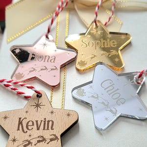 May include: Four star-shaped Christmas ornaments with names: "Fiona", "Sophie", "Chloe", and "Kevin". The ornaments are in various colors: rose gold, gold, silver, and wood. Each has a red and white string for hanging, and a sleigh and reindeer design.