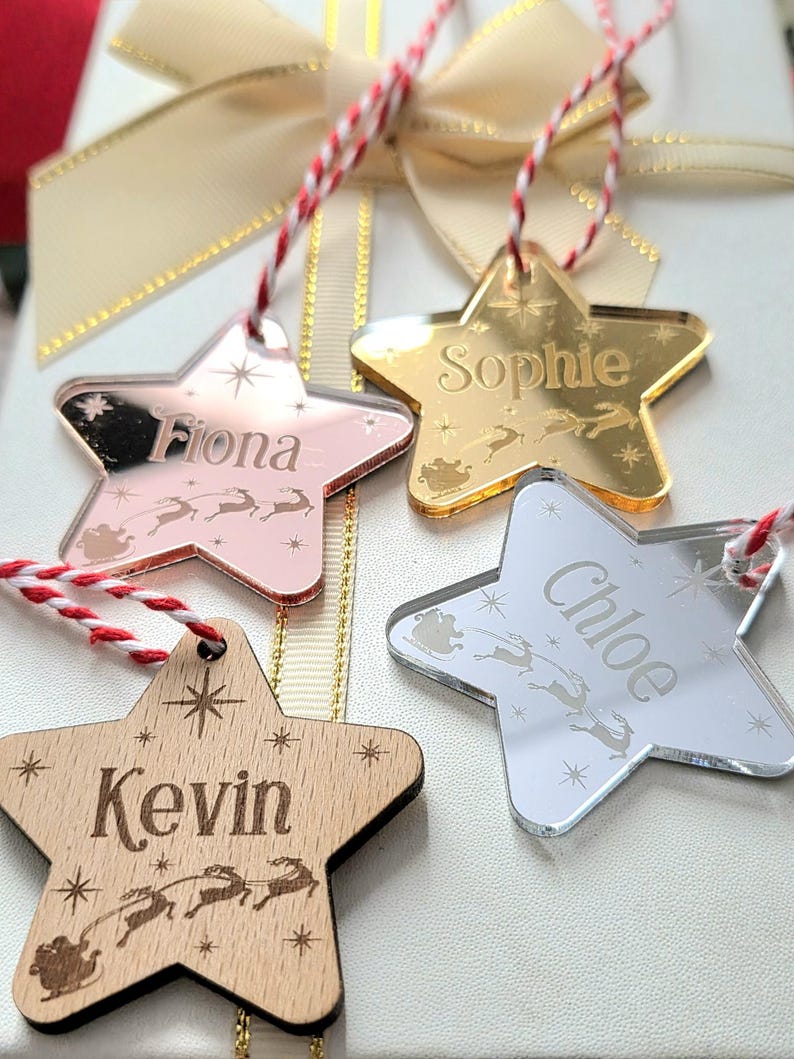 May include: Four star-shaped Christmas ornaments with names: "Fiona", "Sophie", "Chloe", and "Kevin". The ornaments are in various colors: rose gold, gold, silver, and wood. Each has a red and white string for hanging, and a sleigh and reindeer design.