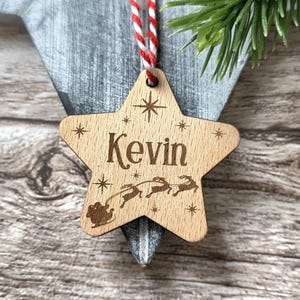 May include: Wooden star-shaped Christmas ornament with the name "Kevin" engraved in a decorative font. The ornament features a sleigh with reindeer and starburst designs. It hangs from a red and white striped string.