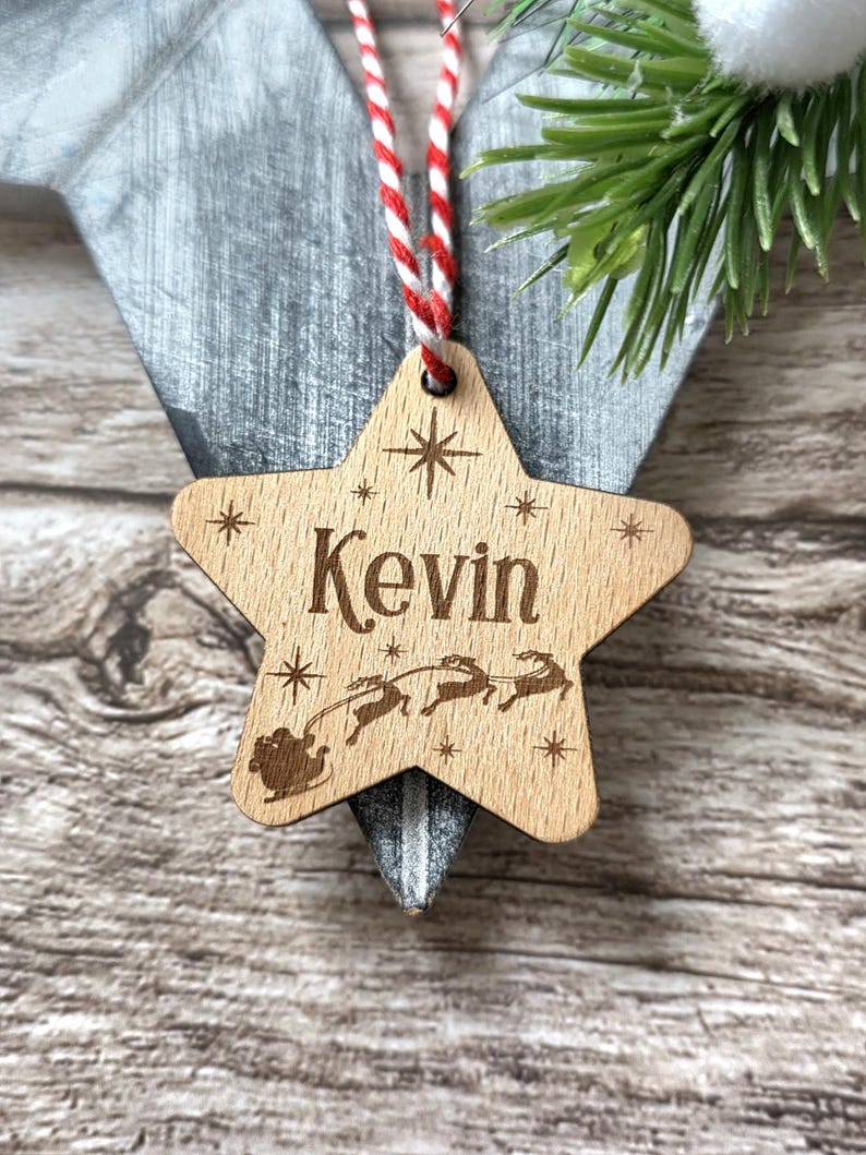 May include: Wooden star-shaped Christmas ornament with the name "Kevin" engraved in a decorative font. The ornament features a sleigh with reindeer and starburst designs. It hangs from a red and white striped string.