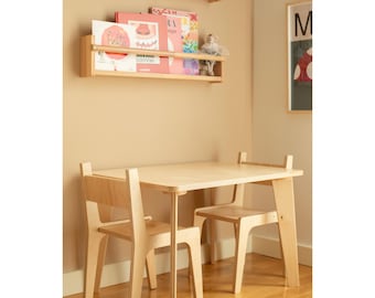May include: A light-colored wooden children's table and two chairs set. The table has a rectangular top and straight legs. The chairs have a simple design with a backrest. The set is in a room with wooden shelves and books.