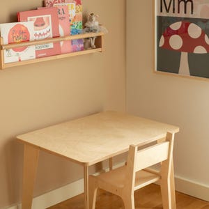 May include: A light wood children's table and chair set. The table is rectangular with rounded corners and angled legs. The chair has a slatted back. The set is in a room with a bookshelf and a mushroom-themed poster.