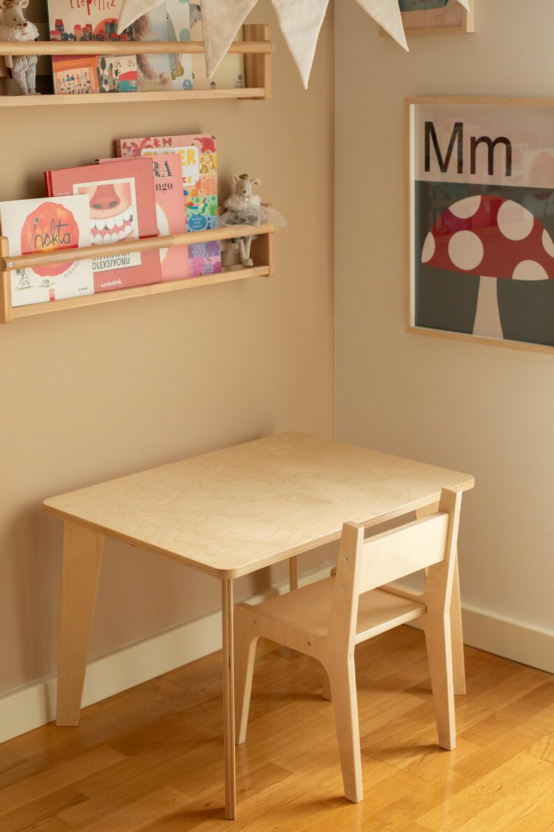 May include: A light wood children's table and chair set. The table is rectangular with rounded corners and angled legs. The chair has a slatted back. The set is in a room with a bookshelf and a mushroom-themed poster.